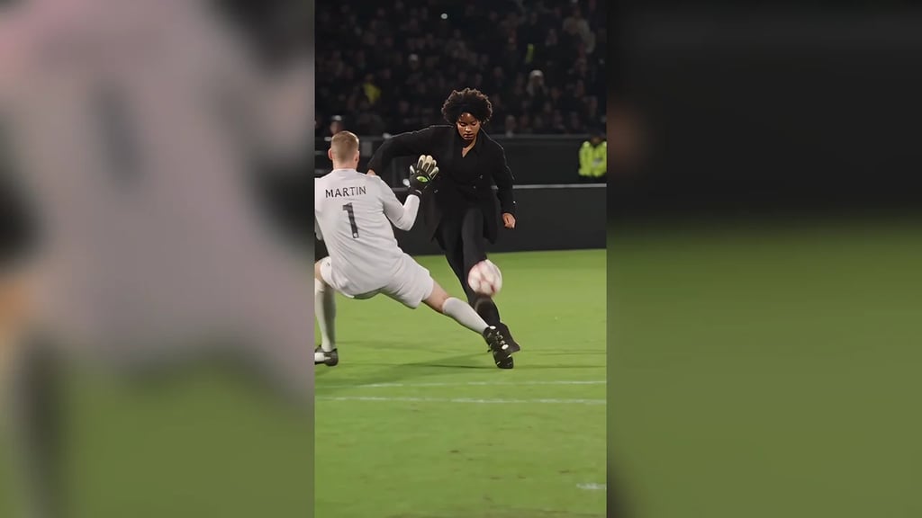 A player in a black suit runs at the diving goalkeeper on a floodlit pitch